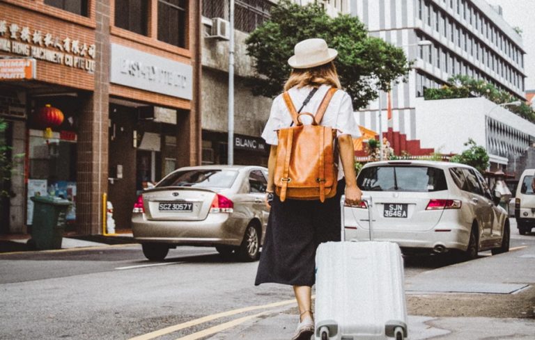 Žena s batohem jdoucí v ulici a v ruce drží kufr.